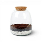 Glass jar with a cork lid containing soil, activated carbon pellets and small gravel stones on a white background