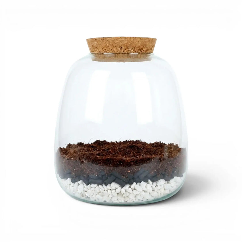 Glass jar with cork lid containing layers of soil, activated carbon pellets, and pebbles on a white background